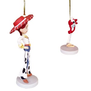 May include: Two Christmas ornaments featuring characters from the Toy Story franchise. The first ornament is a figurine of Jessie, a cowgirl doll with red hair, wearing a red and white hat, a white shirt, and blue jeans with white spots. The second ornament is a figurine of a red and white toy with a blue and white body and a red and white head.