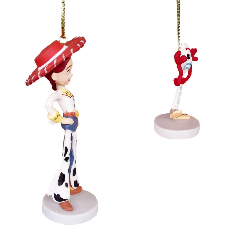 May include: Two Christmas ornaments featuring characters from the Toy Story franchise. The first ornament is a figurine of Jessie, a cowgirl doll with red hair, wearing a red and white hat, a white shirt, and blue jeans with white spots. The second ornament is a figurine of a red and white toy with a blue and white body and a red and white head.