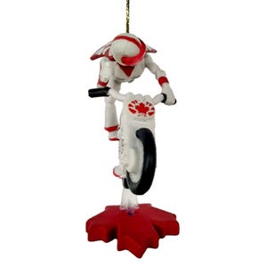 May include: A white and red Christmas ornament of a figure on a motorcycle doing a wheelie. The figure is wearing a helmet and a white and red suit. The motorcycle has a Canadian flag on the side. The ornament is standing on a red maple leaf.
