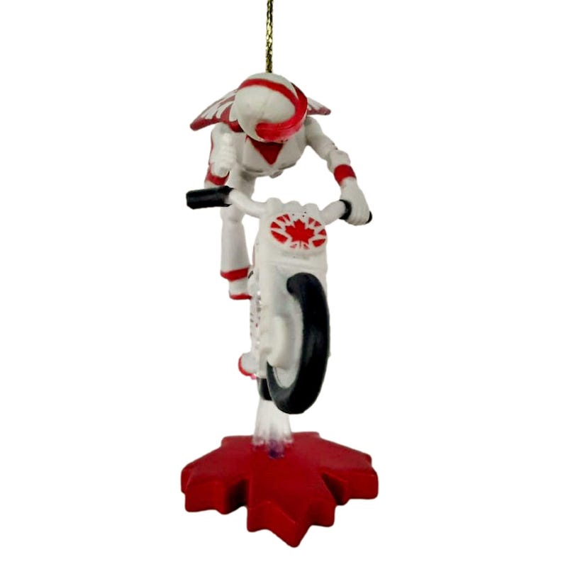 May include: A white and red Christmas ornament of a figure on a motorcycle doing a wheelie. The figure is wearing a helmet and a white and red suit. The motorcycle has a Canadian flag on the side. The ornament is standing on a red maple leaf.