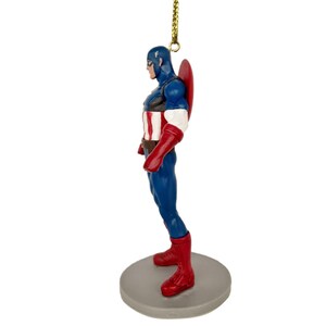 May include: A Captain America figurine in a side profile, featuring a blue and red costume with a shield on the back. The figurine is attached to a gold string for hanging. It stands on a gray circular base.