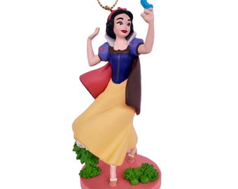 May include: Snow White Christmas ornament featuring a figurine of Snow White in a yellow dress, blue bodice, and red cape, holding a blue bird. The ornament is on a pink base with green foliage.