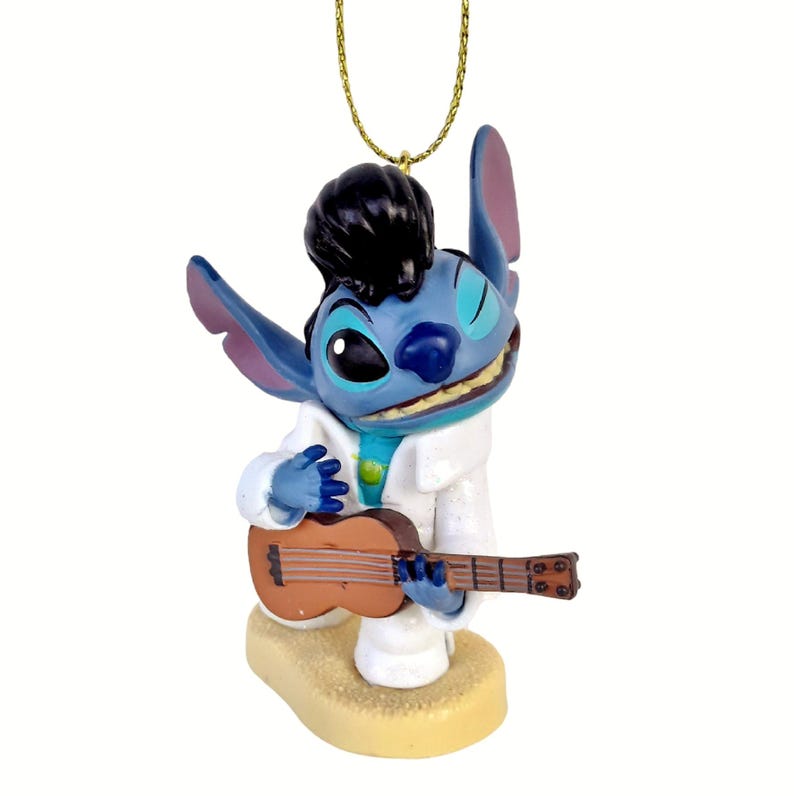 Puede incluir: Una figura azul de Stitch vestido como Elvis Presley, con un traje blanco y sosteniendo un ukelele marr&oacute;n. La figura tiene cabello negro y est&aacute; sonriendo. Est&aacute; sujeta a una cuerda dorada para colgar.