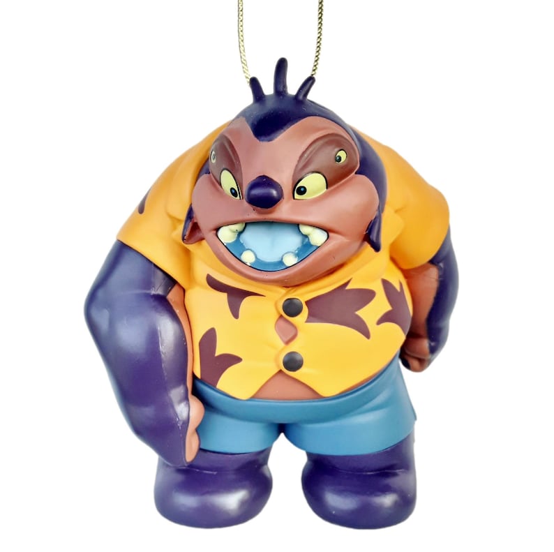 Op de afbeelding: Een beeldje van een stripfiguur met een paarse kop en armen, een oranje shirt en een blauwe korte broek. Het personage heeft een boze uitdrukking met scherpe tanden en een blauwe tong. Bovenop is een gouden touwtje bevestigd.