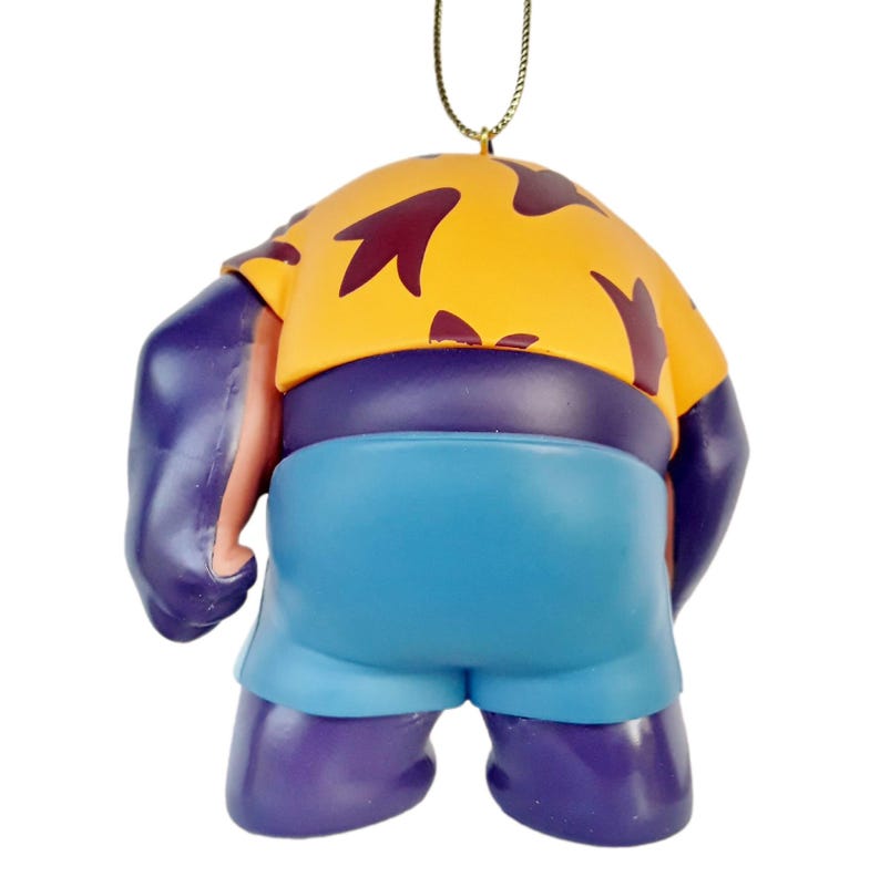 Op de afbeelding: Een paars en blauw beeldje van een stripfiguur, van achteren gezien. Het personage draagt een geel shirt met kastanjebruine bladvormige patronen, een blauwe korte broek en heeft een paarse huid. Een gouden draad is bevestigd om op te hangen.