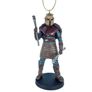 May include: A Christmas ornament featuring a Mandalorian character from Star Wars. The figure is wearing a brown and blue armor with a gold helmet and holding a wrench and a tool. The ornament is on a black base.