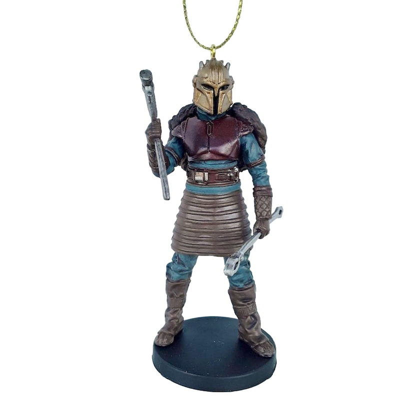 May include: A Christmas ornament featuring a Mandalorian character from Star Wars. The figure is wearing a brown and blue armor with a gold helmet and holding a wrench and a tool. The ornament is on a black base.