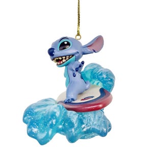 May include: A blue Stitch ornament surfing on a wave. The ornament features Stitch on a surfboard with a red and white stripe. The wave is a translucent blue. The ornament is suspended by a gold string.