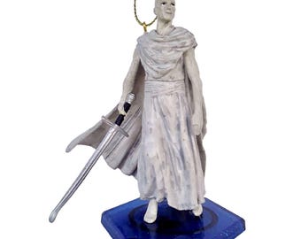 May include: White figurine of a person holding a sword, standing on a blue hexagonal base with glitter. The figurine is wearing a white robe and has a white cape.