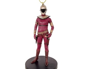 May include: A red and gold action figure ornament of a female character wearing a helmet and holding two blaster pistols. The figure is standing on a black circular base.