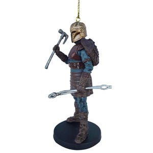 May include: A detailed ornament of a warrior figure with a gold helmet, brown armor, and a blue tunic. The figure is holding a silver hammer in one hand and a silver tool in the other. The ornament is standing on a black base.