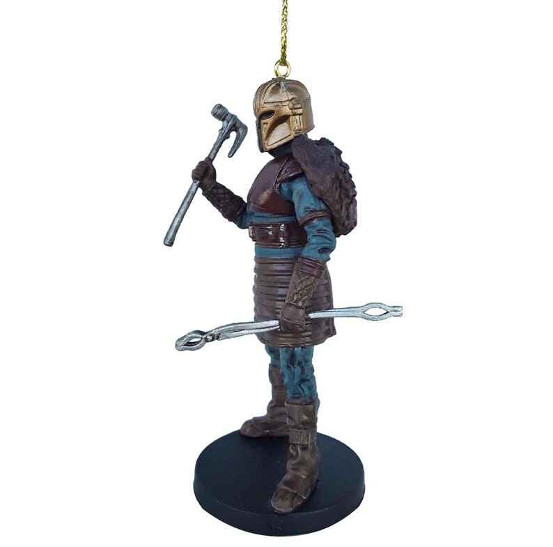 May include: A detailed ornament of a warrior figure with a gold helmet, brown armor, and a blue tunic. The figure is holding a silver hammer in one hand and a silver tool in the other. The ornament is standing on a black base.
