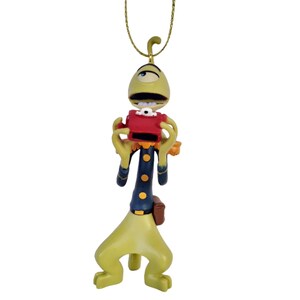 May include: A whimsical ornament of a cartoon alien figure. The alien is primarily yellow-green with a red camera and a dark blue uniform with gold buttons. It hangs from a gold-colored chain, perfect for holiday decor.