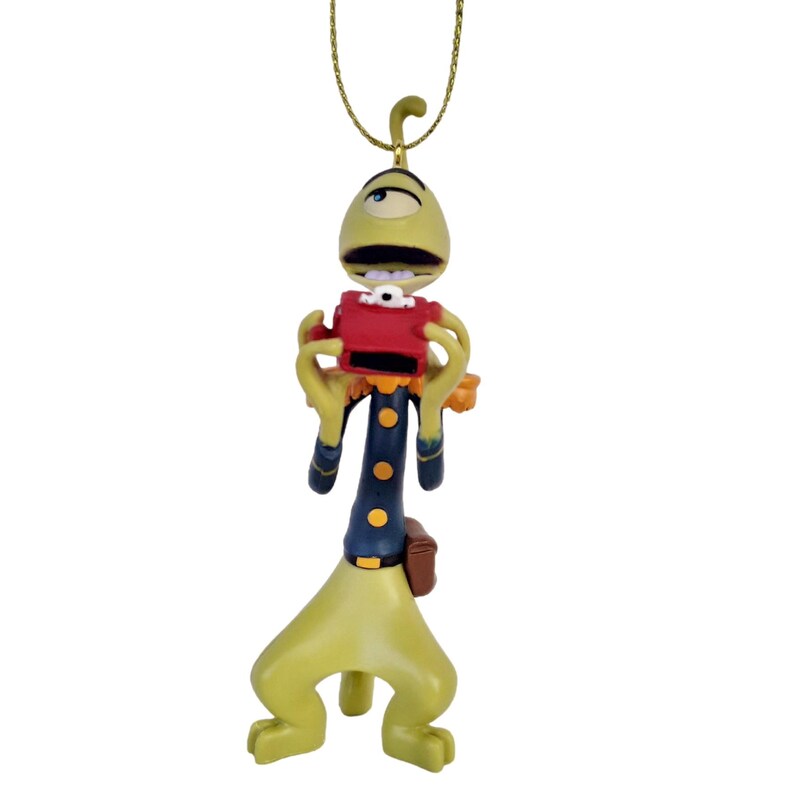May include: A whimsical ornament of a cartoon alien figure. The alien is primarily yellow-green with a red camera and a dark blue uniform with gold buttons. It hangs from a gold-colored chain, perfect for holiday decor.