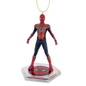 May include: A Spider-Man figure in a standing pose, featuring a red and gold suit with black accents. The figure is attached to a gold-colored string and sits on a clear, hexagonal base with a red spider-web design.