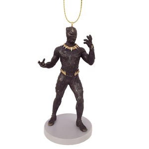 May include: A Black Panther figurine with gold accents, standing on a gray base. The superhero figure has a gold necklace and belt, and is suspended by a gold string. The figurine is detailed and appears to be made of a dark, possibly metallic material.