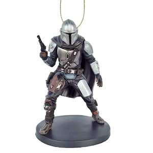 May include: A silver and black Mandalorian action figure ornament with a blaster pistol in his right hand. He is wearing a cape and standing on a black base.