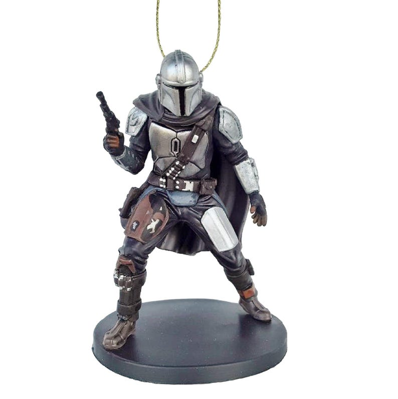 May include: A silver and black Mandalorian action figure ornament with a blaster pistol in his right hand. He is wearing a cape and standing on a black base.