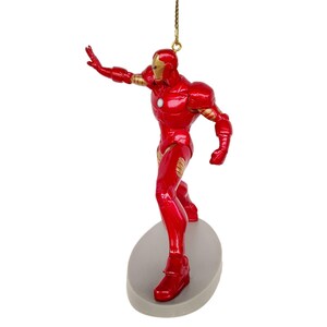 May include: A red and gold Iron Man figurine ornament. The superhero figure is posed with one arm outstretched, mounted on a gray oval base. A gold string is attached for hanging. The ornament is a collectible item.