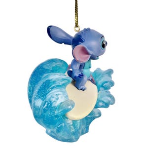 May include: A blue Stitch figurine riding a surfboard on a wave. The figurine has large ears, a pink nose, and a small blue body. The wave is a translucent blue with white accents, and the surfboard is off-white.