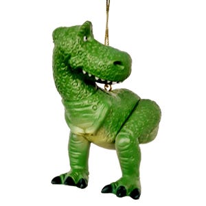 Puede incluir: Adorno de pl&aacute;stico verde de un dinosaurio de la franquicia de pel&iacute;culas Toy Story. El dinosaurio est&aacute; de pie sobre sus cuatro patas con la cabeza girada hacia un lado. El adorno tiene una cuerda dorada para colgar.