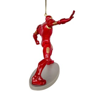 May include: A red Iron Man figurine with gold accents, posed as if surfing on a gray oval base. The figurine is suspended by a gold string, suggesting it's an ornament. The figure is in a dynamic pose, with one arm outstretched.