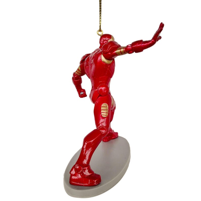 May include: A red Iron Man figurine with gold accents, posed as if surfing on a gray oval base. The figurine is suspended by a gold string, suggesting it's an ornament. The figure is in a dynamic pose, with one arm outstretched.