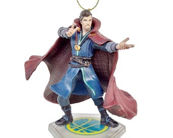 May include: A Doctor Strange figurine ornament. The figure is in a dynamic pose, wearing a blue and red costume with a cape. The ornament has a gold string for hanging and is on a clear, hexagonal base with a gold and blue design.