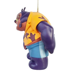 Op de afbeelding: Een beeldje van een stripfiguur, met een paars lichaam, bruin gezicht en geel shirt met een donkerpaars ontwerp. Het personage draagt een blauwe korte broek. Het beeldje hangt aan een gouden touwtje.