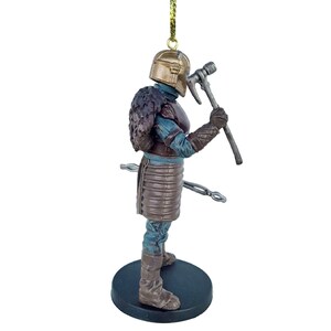 May include: A brown and gold ornament of a warrior figure with a helmet, fur, and a weapon. The figure is standing on a black base.