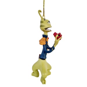 May include: A whimsical alien Christmas ornament. The figure is light green with dark green stripes, wearing a blue jacket with gold accents and holding a red object. The ornament hangs from a gold string.