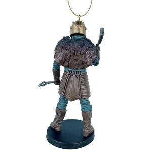 May include: A brown and blue figurine of a warrior wearing a fur-lined helmet and holding a weapon. The figurine is standing on a black base and has a gold string attached to the top of the helmet.
