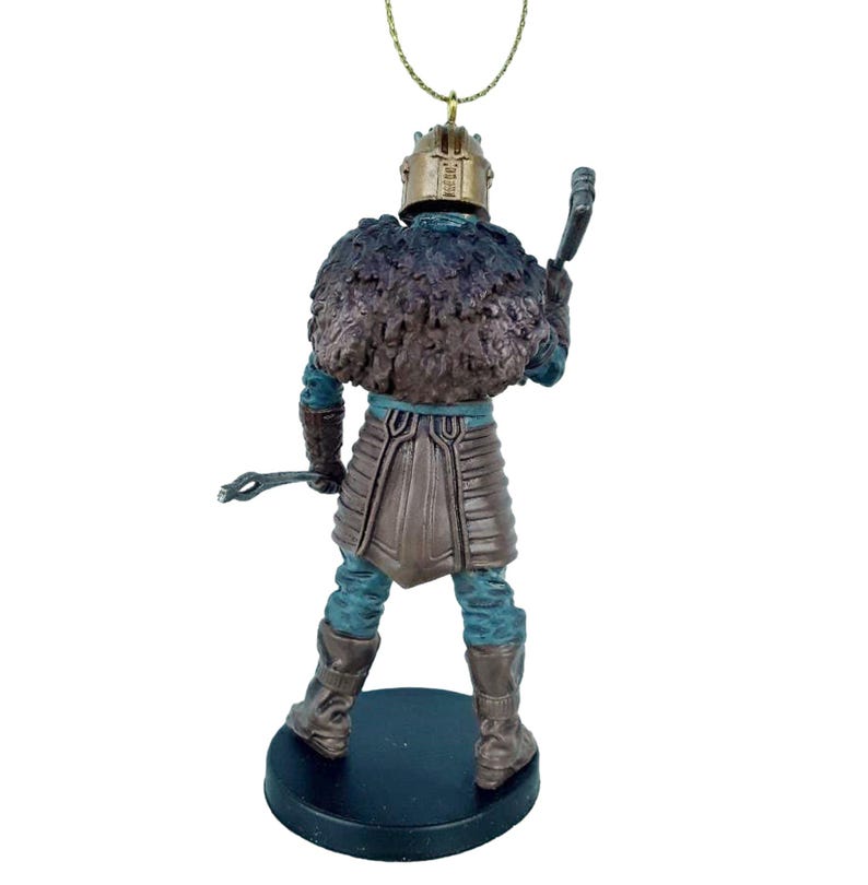 May include: A brown and blue figurine of a warrior wearing a fur-lined helmet and holding a weapon. The figurine is standing on a black base and has a gold string attached to the top of the helmet.