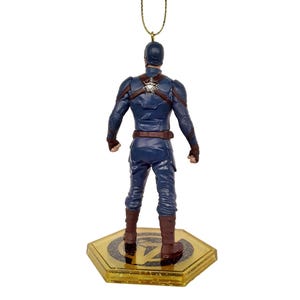 May include: A Captain America figurine, viewed from the back, stands on a gold-colored hexagonal base. The figurine is dressed in a blue suit with brown accents and boots. A gold string is attached to the top of the figurine for hanging.