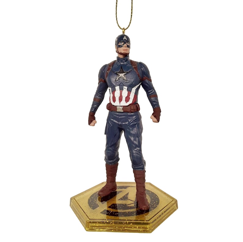 May include: A Captain America figurine ornament. The figurine is in a standing pose, wearing a blue and red costume with a white star on the chest. The ornament is attached to a gold string and sits on a gold base with the Avengers logo.