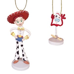May include: Two Christmas ornaments featuring characters from the Toy Story franchise. The first ornament is a red-haired female character wearing a red and white cowboy hat, a white shirt with yellow accents, blue jeans, and black and white cow print pants. The second ornament is a red, blue, and yellow character with a fork-shaped head and a surprised expression.