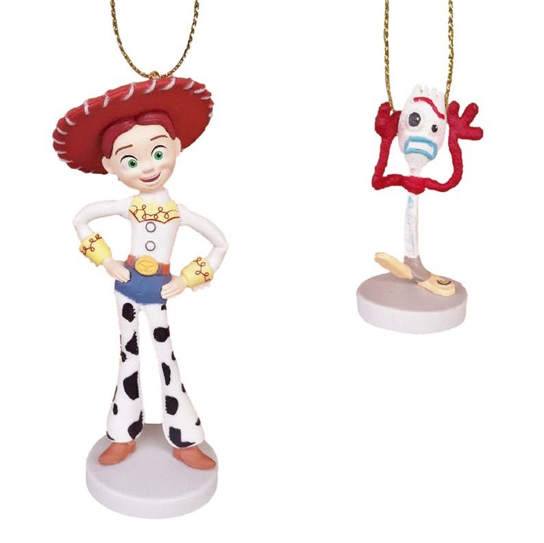 May include: Two Christmas ornaments featuring characters from the Toy Story franchise. The first ornament is a red-haired female character wearing a red and white cowboy hat, a white shirt with yellow accents, blue jeans, and black and white cow print pants. The second ornament is a red, blue, and yellow character with a fork-shaped head and a surprised expression.