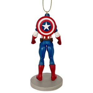 May include: A Captain America figurine ornament, viewed from the back. The figure is in a classic superhero pose, wearing a blue suit, red boots, and a shield with a white star on a red and white background. The ornament hangs from a gold string.
