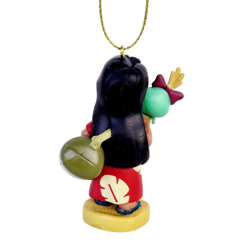 Pode incluir: Estatueta de uma personagem de um filme de anima&ccedil;&atilde;o popular, vista por tr&aacute;s. A figura tem cabelo preto, um vestido vermelho com um desenho branco e segura um objeto verde. A estatueta est&aacute; sobre uma base amarela e tem um fio dourado para pendurar.