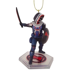 May include: A Taskmaster figure ornament, featuring a character in a blue, red, and white costume, holding a sword and shield. The figure stands on a clear, hexagonal base with a red emblem, suspended by a gold string.