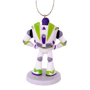 May include: A white, green, and purple plastic figurine of Buzz Lightyear from the Toy Story franchise. The figurine is standing with its arms crossed and is wearing a space ranger suit. The figurine is attached to a gold string, making it a Christmas ornament.