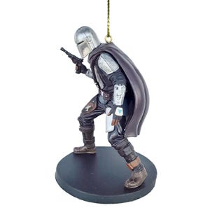 May include: A silver and black Mandalorian ornament with a cape, holding a blaster pistol. The ornament is standing on a brown base.