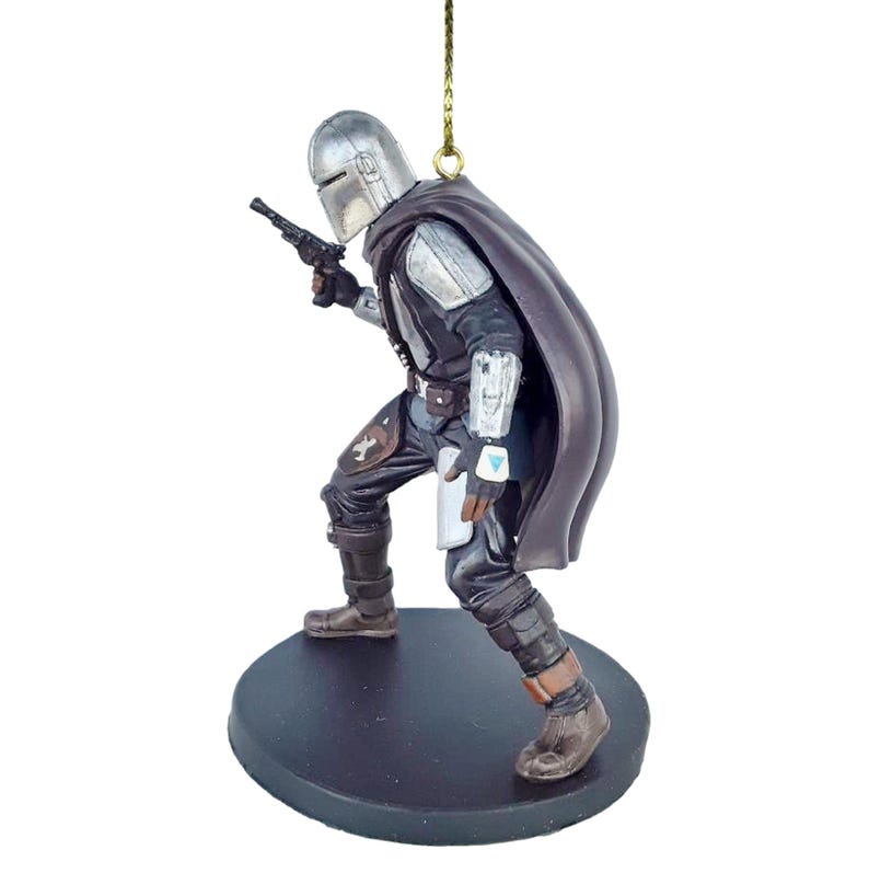 May include: A silver and black Mandalorian ornament with a cape, holding a blaster pistol. The ornament is standing on a brown base.