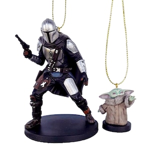 May include: Two detailed resin ornaments featuring characters from a popular science fiction series. One depicts a figure in silver armour holding a blaster, the other a small green creature in a brown robe. Both have gold hanging cords.