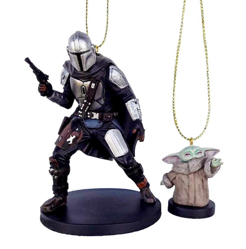May include: Two detailed resin ornaments featuring characters from a popular science fiction series. One depicts a figure in silver armour holding a blaster, the other a small green creature in a brown robe. Both have gold hanging cords.