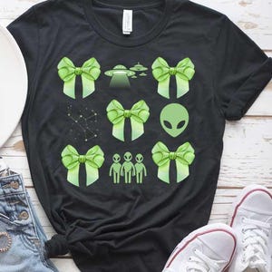 May include: Black t-shirt featuring a green alien, green bows, and UFOs printed on it. The shirt is paired with denim shorts and white trainers.