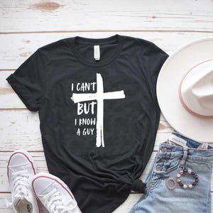 May include: Black t-shirt with a white cross and the text "I can't but I know a guy". The shirt is paired with denim shorts, a white hat, and white sneakers.