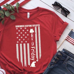 Può includere: Maglietta rossa con un designo di bandiera americana bianca e il testo "Remember Everyone Deployed Red Friday". La maglietta è su una superficie di legno bianco con un paio di scarpe da ginnastica bianche, un paio di occhiali da sole e un paio di jeans blu.