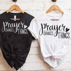 Peut inclure: Deux t-shirts avec le texte "prayer changes things" et un petit graphique de cœur. Un t-shirt est noir et l'autre est blanc.