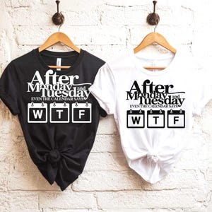 Könnte beinhalten: Zwei T-Shirts, eines schwarz und eines weiß, hängen an Holzbügeln. Jedes Shirt trägt den Text "After Monday and Tuesday, even the calendar says WTF" in Weiß bzw. Schwarz. Die Shirts werden vor einer weißen Backsteinmauer präsentiert.